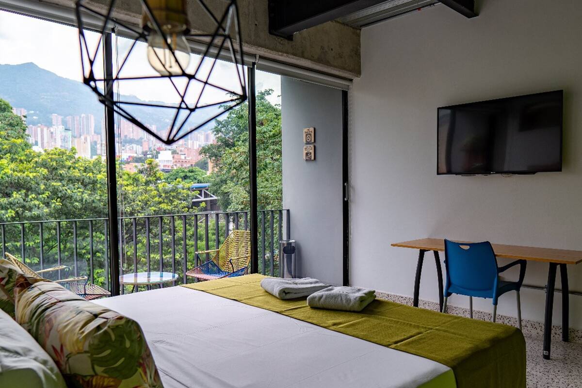 A bedroom with a bed, a desk, and a chair. There is a TV on the wall and a window with a view of buildings and trees.