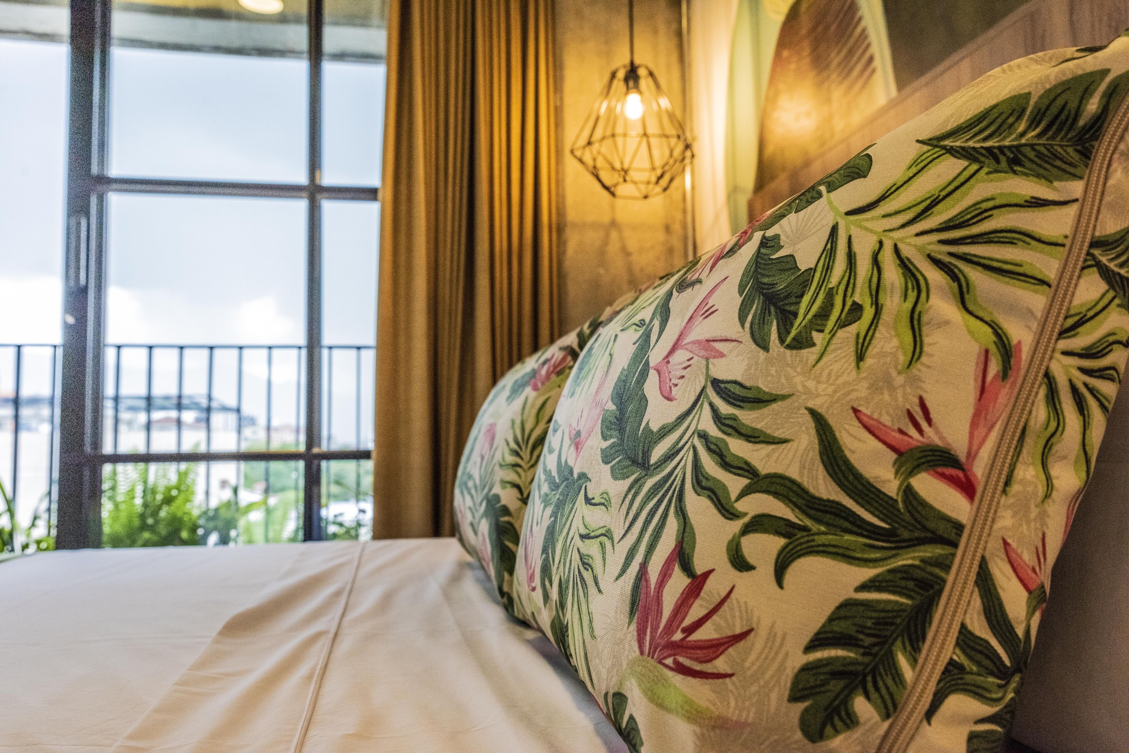 A bed with floral-patterned pillows, a view of greenery outside, and a hanging light fixture.