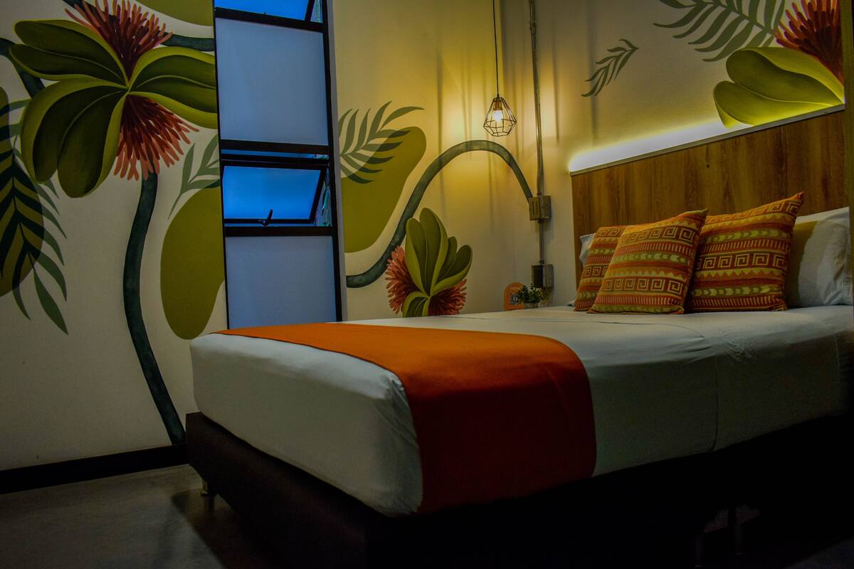 A hotel room with a bed, a nightstand, a wall with a plant mural, and a hanging light fixture.
