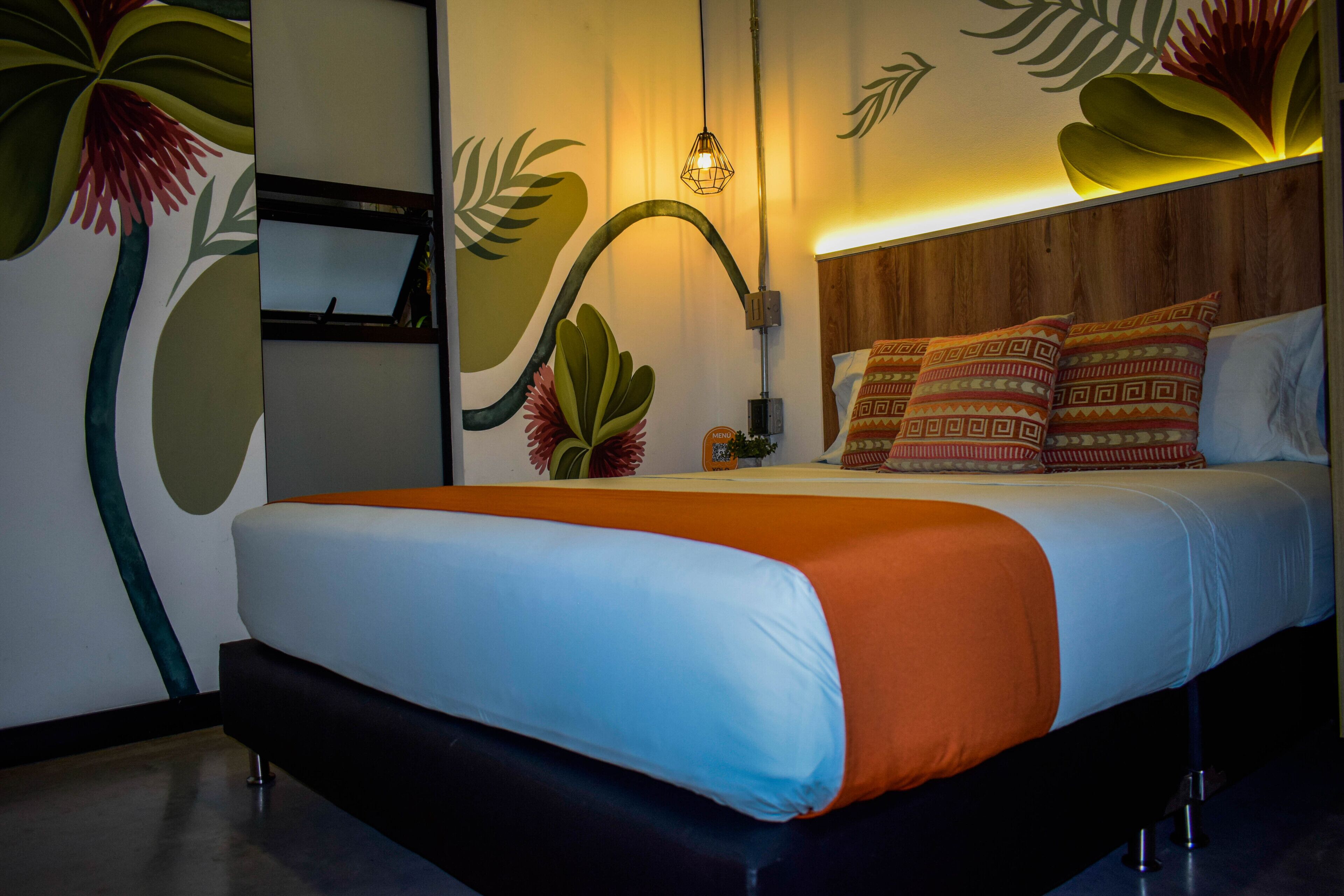 Ein modernes Hotelzimmer mit einem Bett, einem Holz-Kopfschild und einer Wandmalerei mit tropischen Blättern.