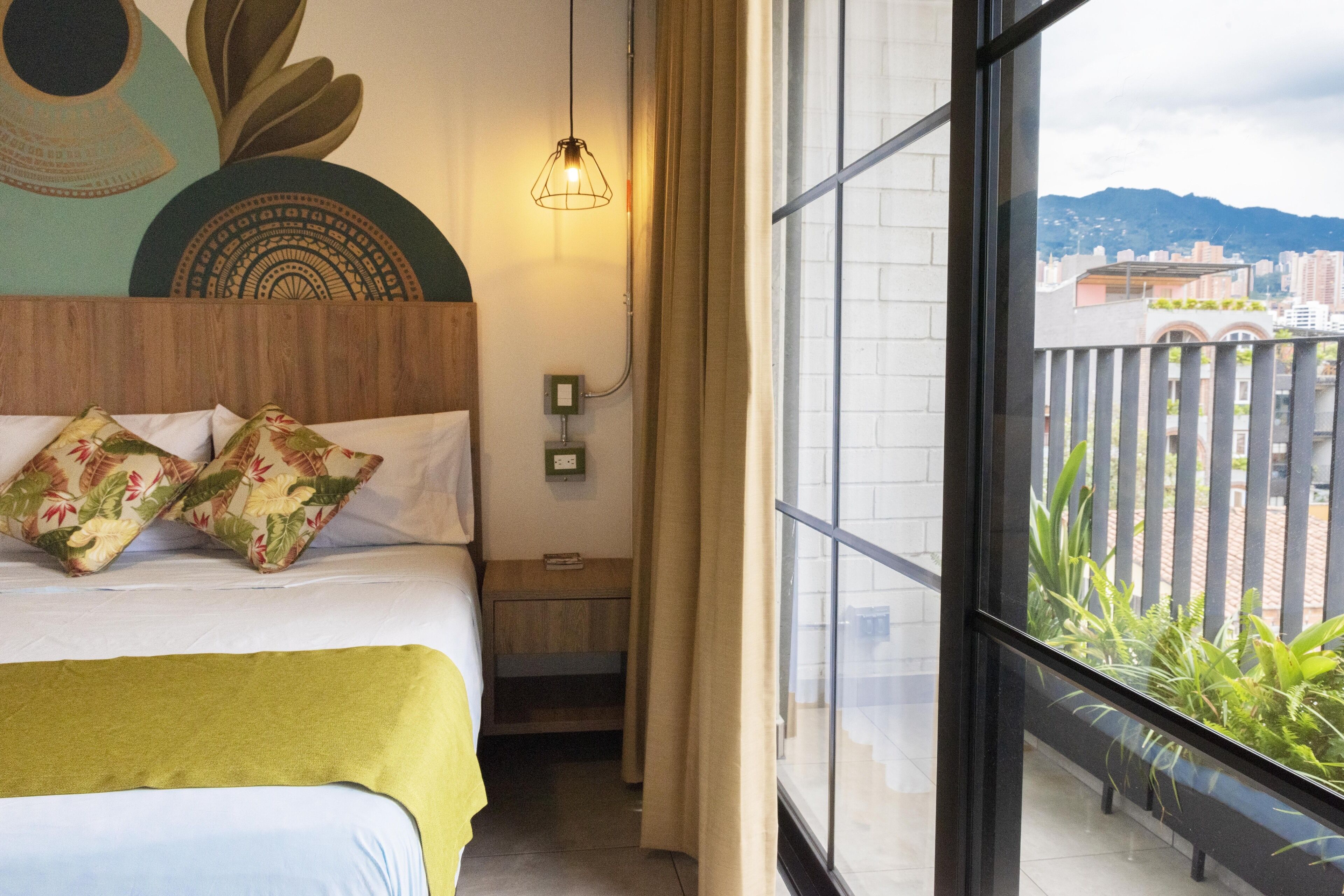 A hotel room with a bed, a green throw blanket, floral pillows, a wall art piece, a hanging light fixture, and a view of a cityscape through a balcony.