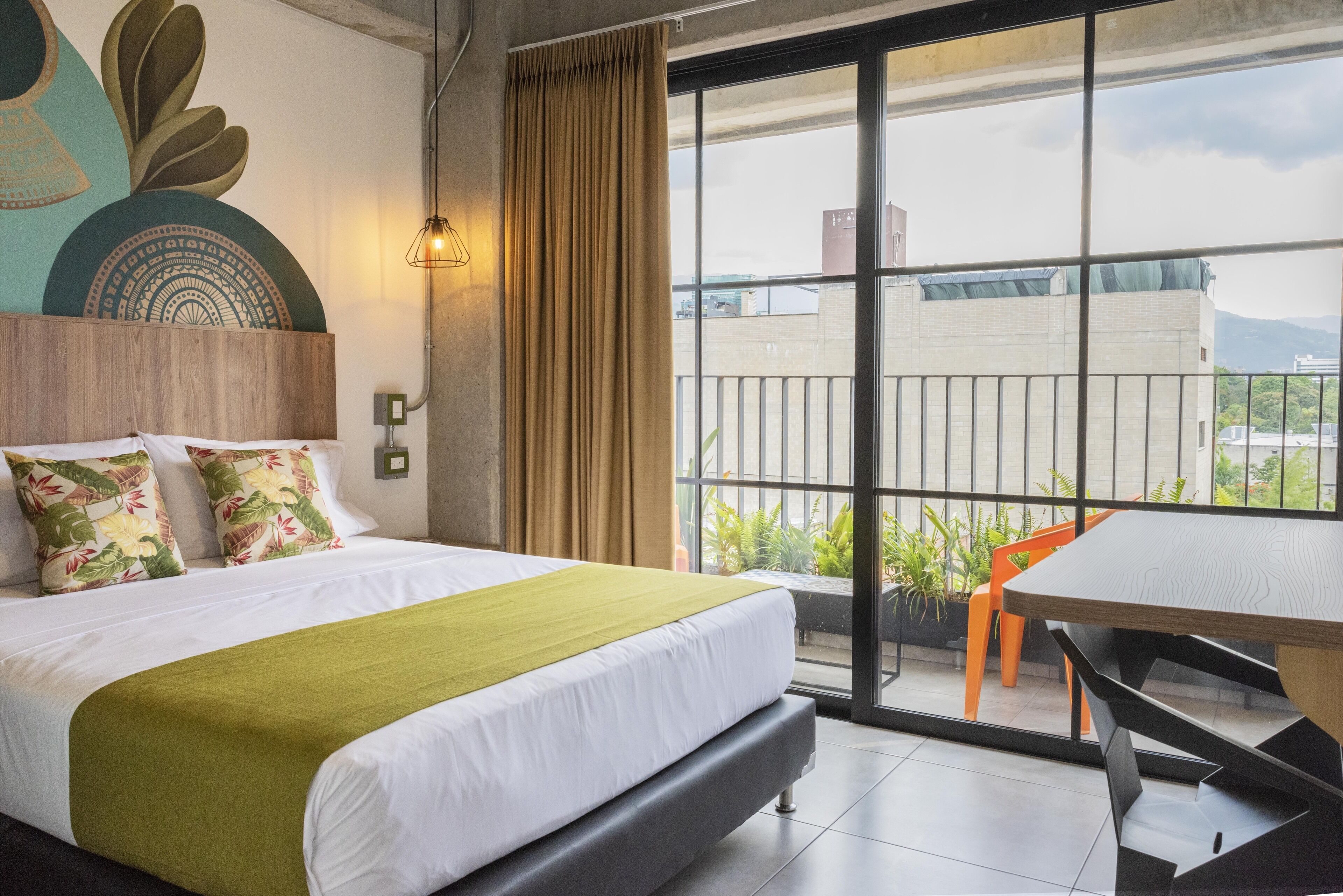 Habitación de hotel con una cama grande, un balcón con sillas naranjas y vistas a edificios y montañas.