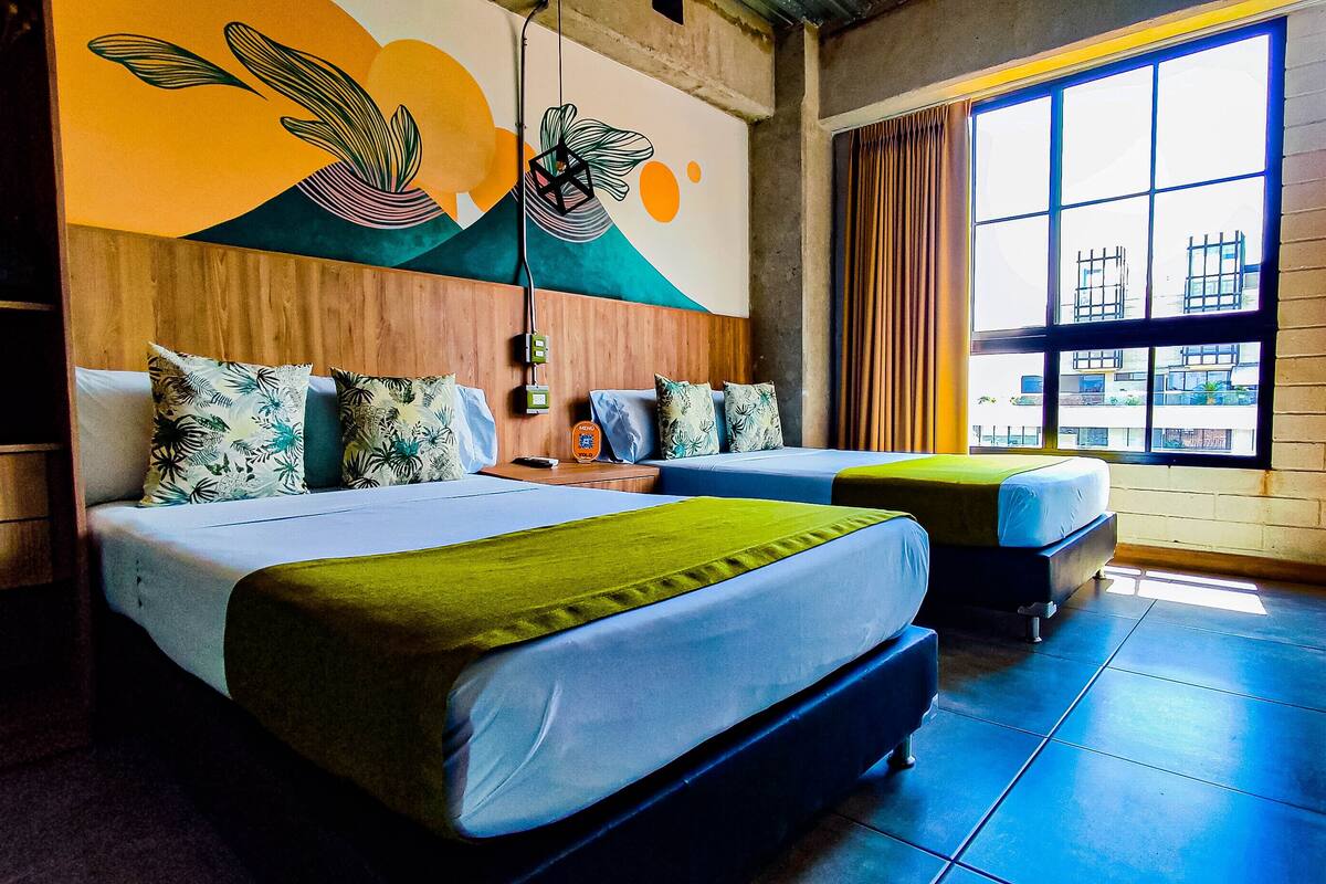 A hotel room with two beds, a wooden headboard, a large window with curtains, and a colorful mural on the wall.