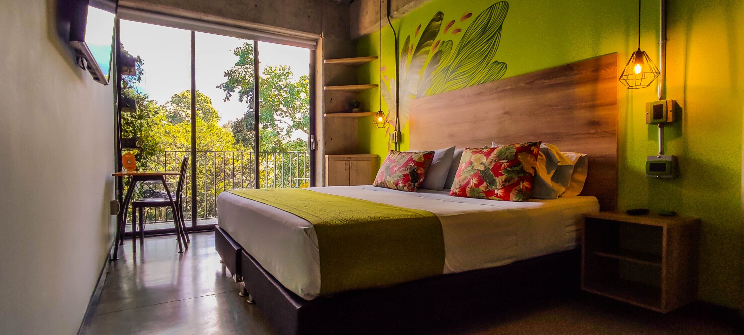 A bedroom with a bed, a nightstand, a chair, a window with a view of trees, and a green wall with a leaf design.