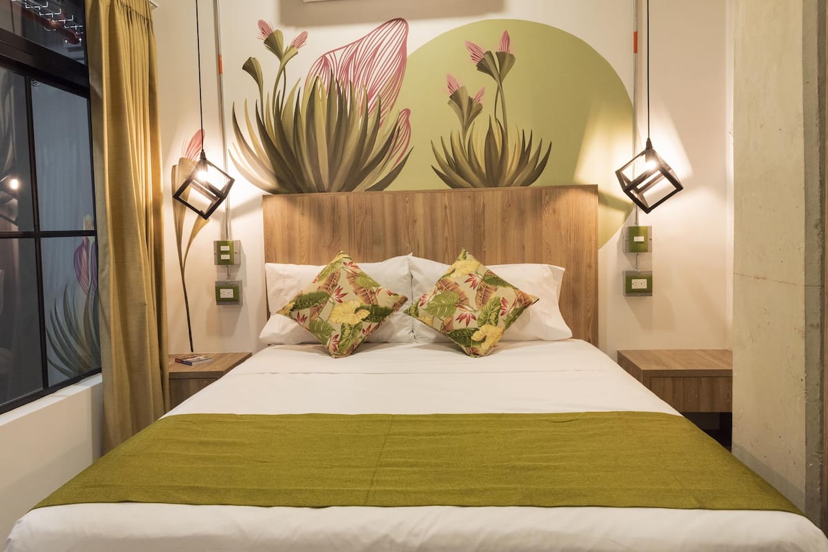 A hotel room with a bed, two nightstands, a green throw blanket, and a wall mural of plants.