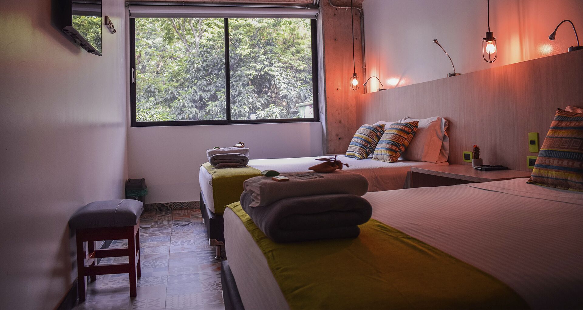 A bedroom with two beds, a bench, a window with a view of trees, and a wall-mounted lamp.
