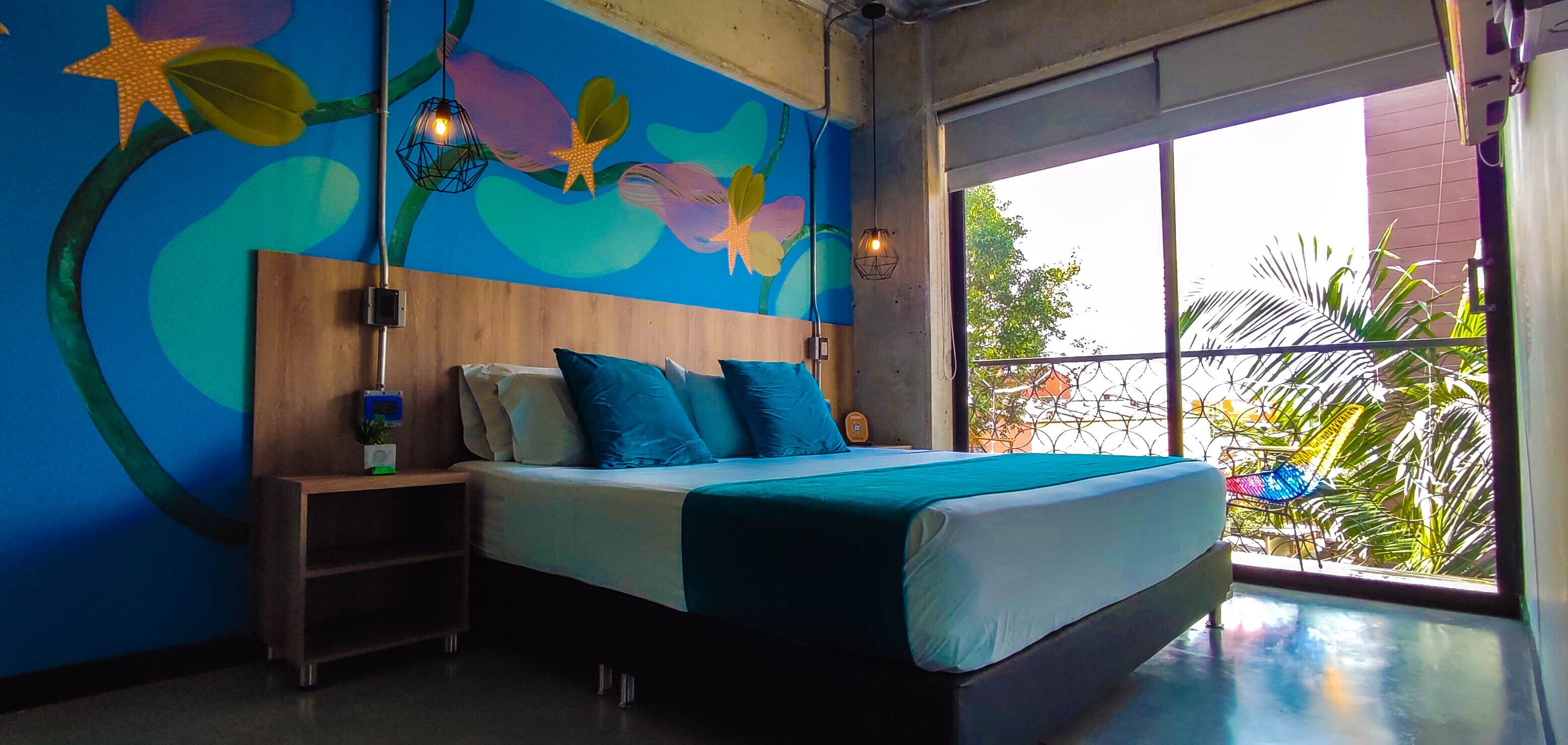 A bedroom with a large bed, a nightstand, a wall mural, and a view of the outdoors.