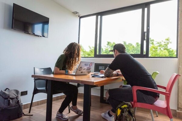 Dos personas sentadas a una mesa con computadoras portátiles, un televisor montado en la pared y una ventana con vistas a árboles.