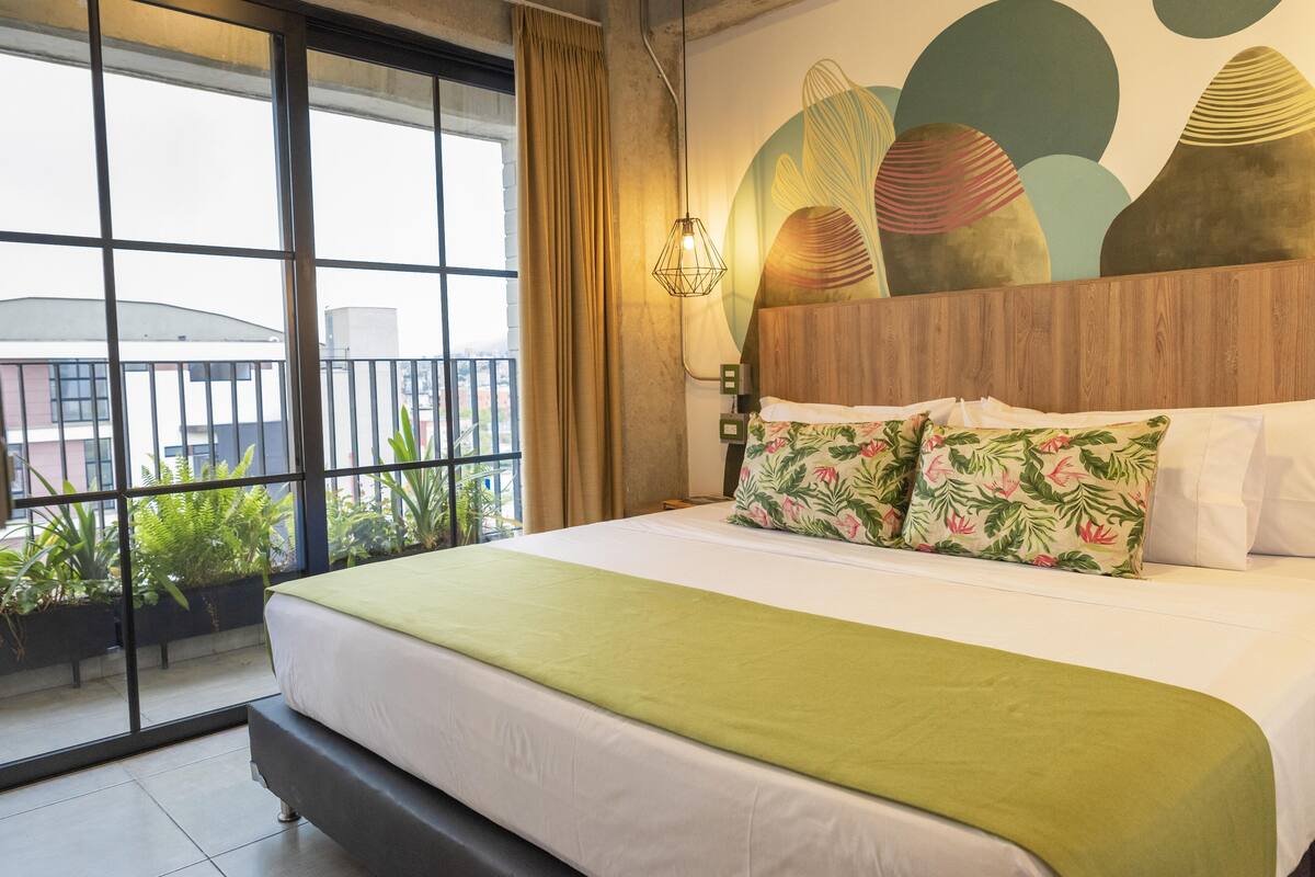 A hotel room with a large bed, a view through a window, and a decorative wall.