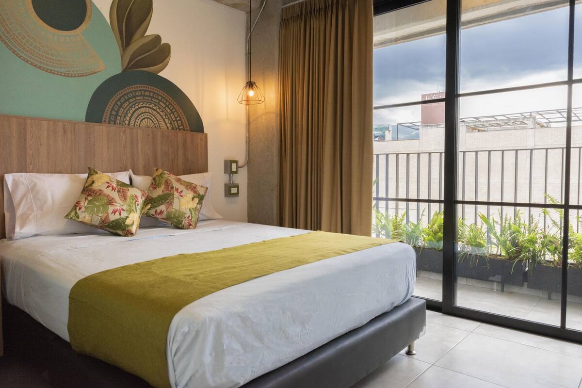 A modern hotel room with a large bed, a wooden headboard, a green throw blanket, and a view of the city through large windows.