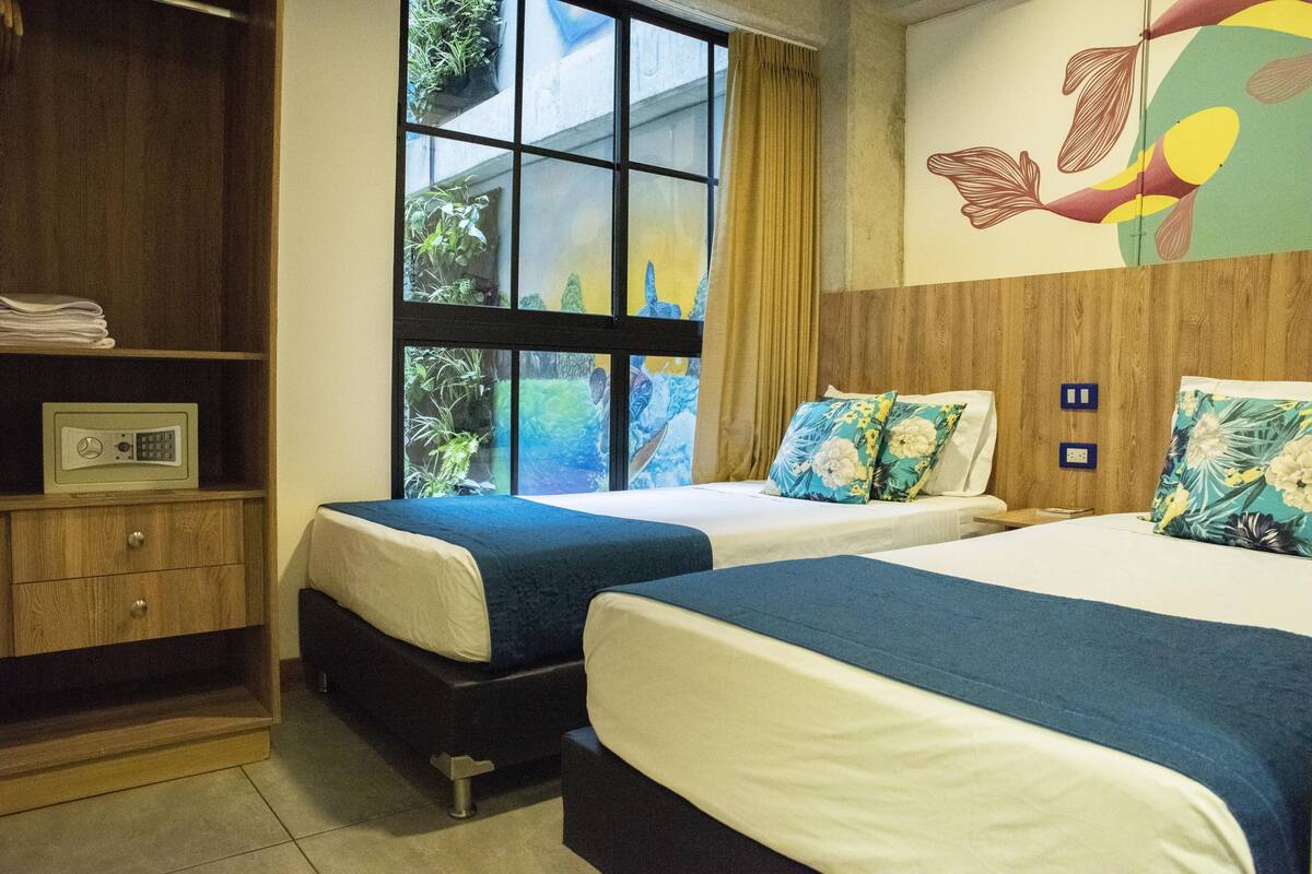 A hotel room with two beds, a wooden wardrobe, a window with a view of greenery, and a wall with a colorful mural.