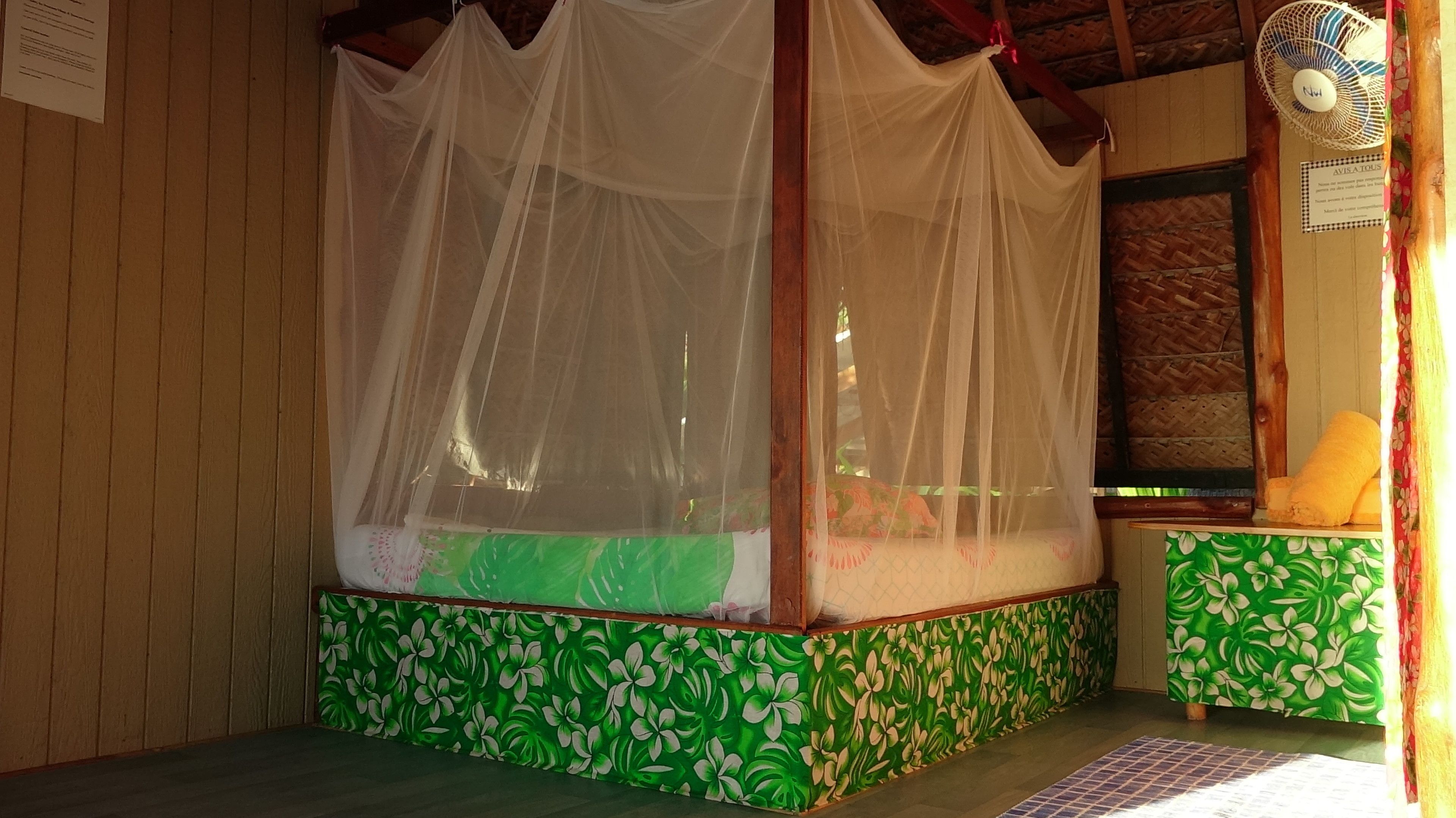 A room with a bed under a mosquito net, green floral wallpaper, a wooden ceiling, and a wall-mounted fan.