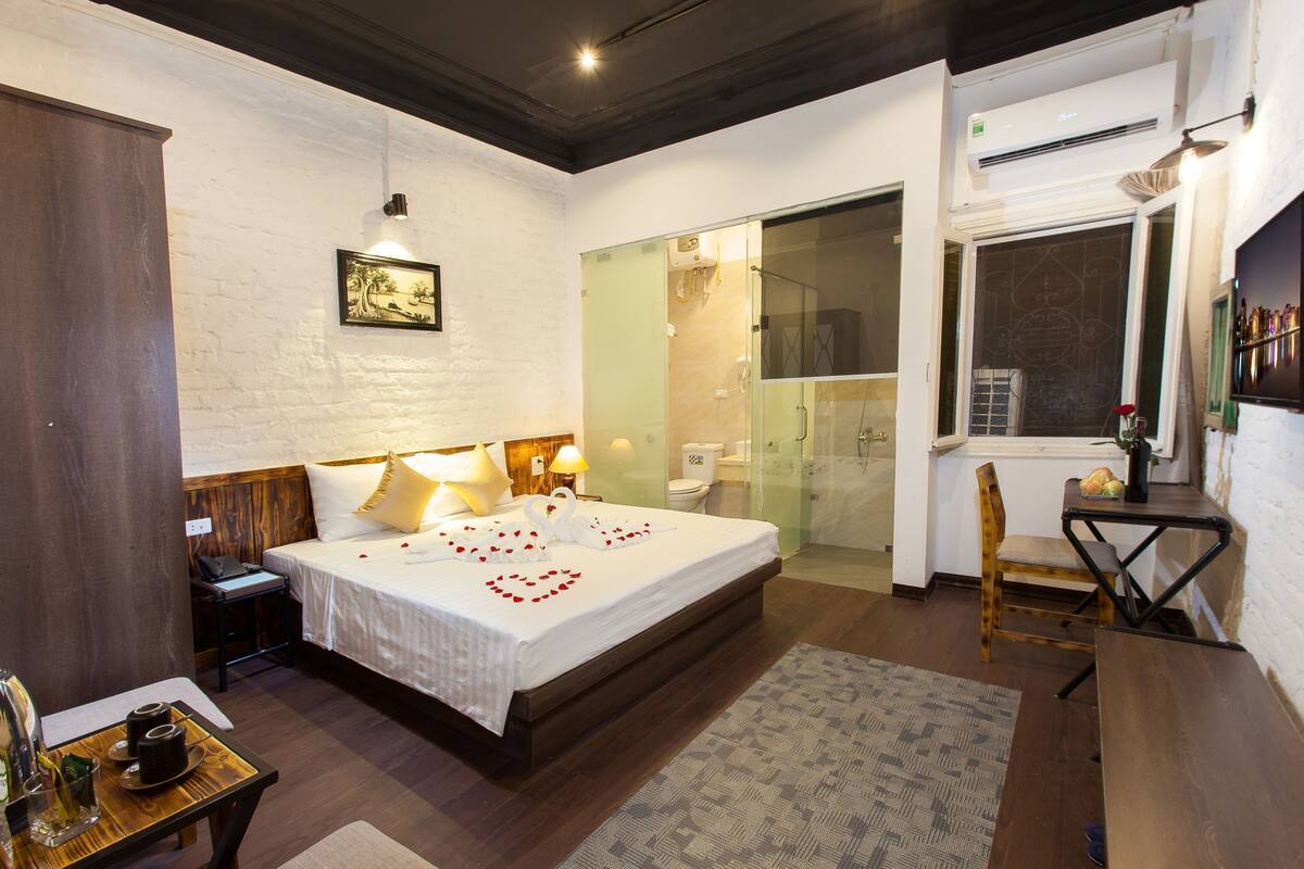 A hotel room with a large bed, a desk with a chair, a small table with drinks, and a bathroom with a glass shower enclosure.