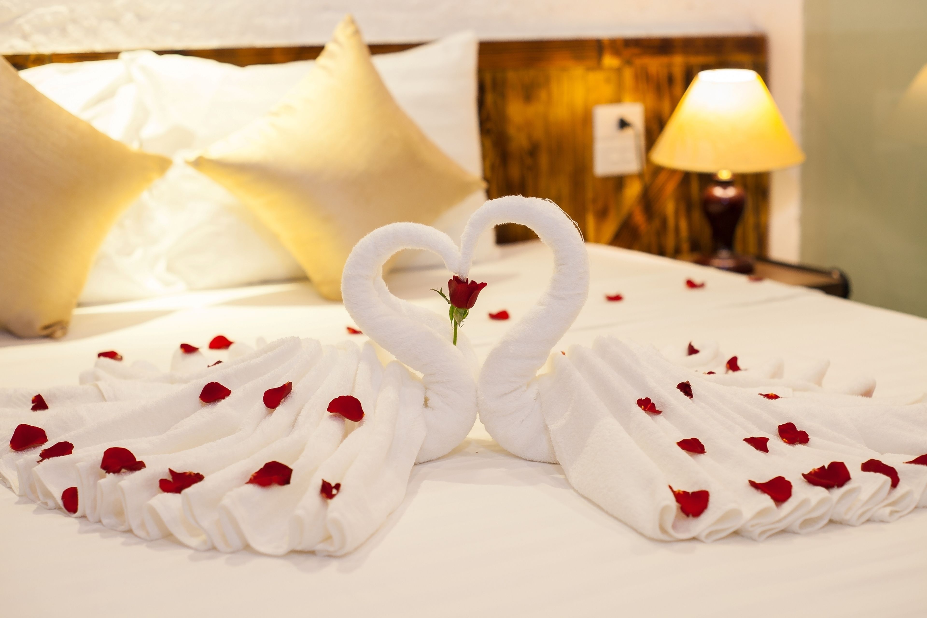 A bed with white linens, a heart-shaped towel arrangement, and rose petals.