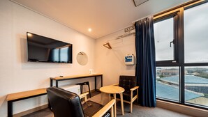 Ein Hotelzimmer mit Flachbildfernseher, Schreibtisch, Stuhl, kleinem Tisch und großem Fenster mit Blick auf Gebäude.