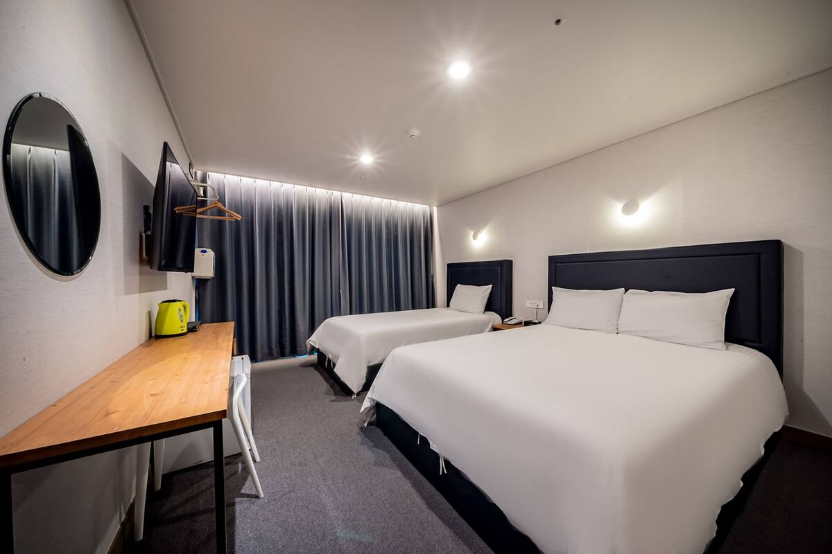 Habitación de hotel con dos camas individuales, un escritorio de madera, un espejo, un perchero y una televisión de pantalla plana.