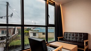 Ein Zimmer mit großem Fenster, einer schwarzen Ledersofa, einem Holzstuhl und einem kleinen Tisch.