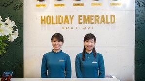 A reception desk with clocks displaying times from New York, London, Hanoi, Paris, and Seoul. The wall behind has a floral pattern and the name "HOLIDAY EMERALD BOUTIQUE" is visible. Two staff members are standing at the desk. - Holiday Emerald Hotel (Hanoi)