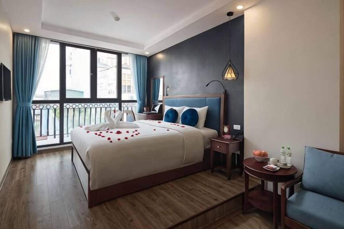 Une chambre d’hôtel avec un grand lit, une table de chevet, une petite table ronde, un fauteuil bleu et un balcon avec des rideaux.
