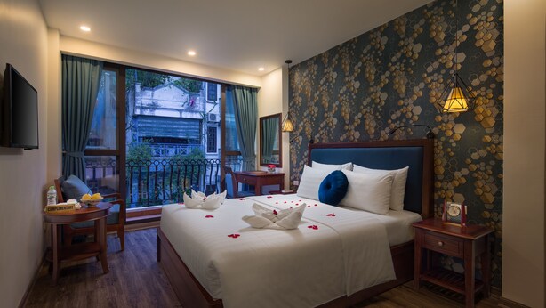 A hotel room with a large bed, a bedside table, a chair, a small table with a bottle and fruit, a TV, and a balcony with curtains. - Holiday Emerald Hotel (Hanoi)