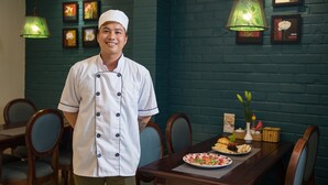 A chef in a white uniform standing in a restaurant with a wooden table set for a meal, framed pictures on the wall, and hanging green lamps. - Holiday Emerald Hotel (Hanoi)