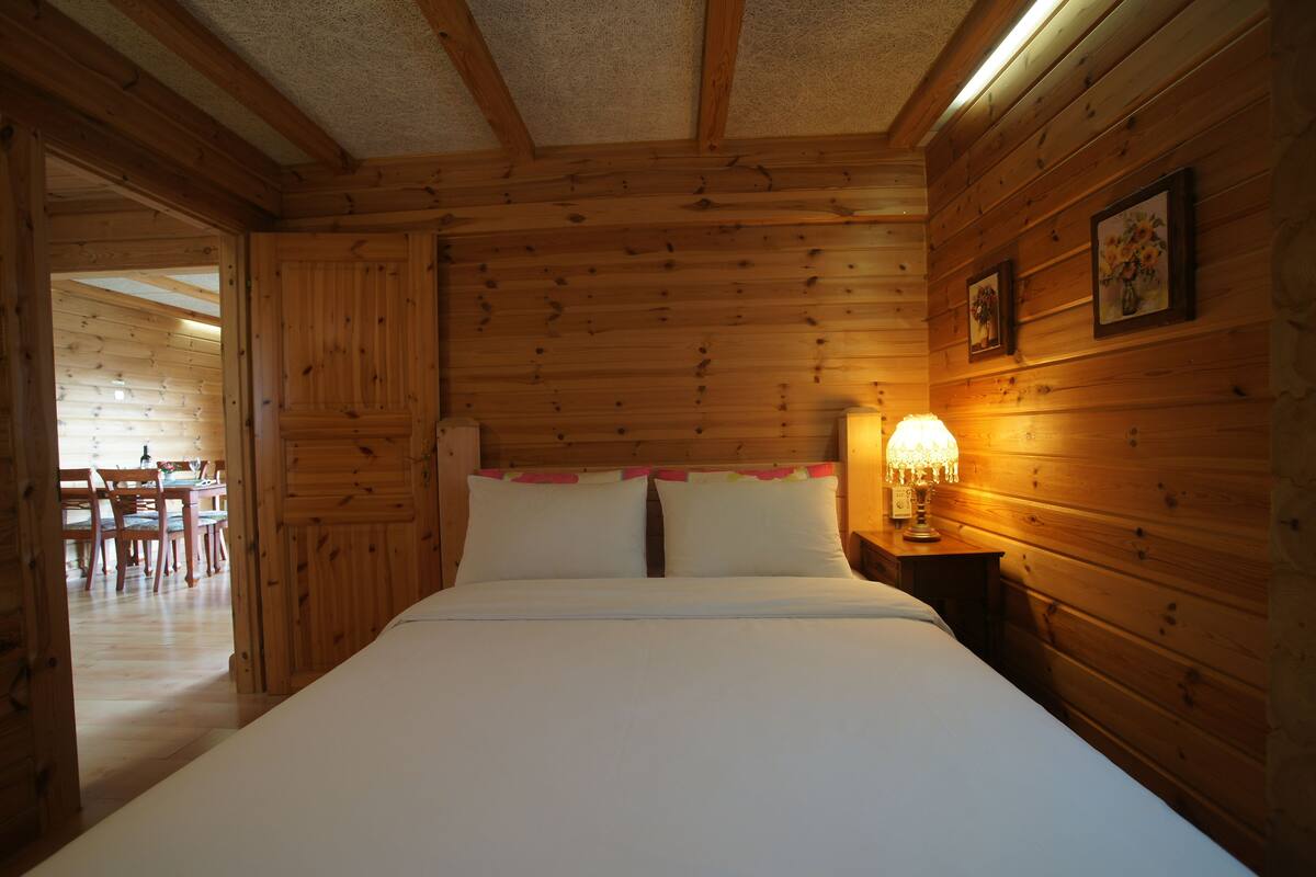 Ein Schlafzimmer einer Holzhütte mit einem Bett, einem Nachttisch mit Lampe und einem Essbereich, der durch eine offene Tür sichtbar ist.