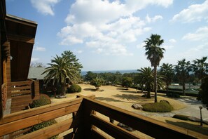 A wooden deck with a view of a landscaped area including palm trees and a distant body of water. - Sunnyday Jeju (Seogwipo)