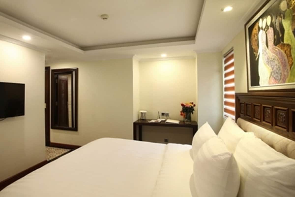 Ein Hotelzimmer mit einem großen Bett, einem flachbildfernseher an der Wand, einem Schreibtisch mit Blumen und einem Gemälde darüber.