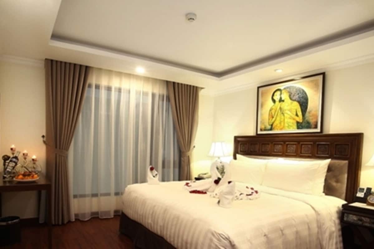 Ein Hotelzimmer mit einem großen Bett, einem Nachttisch, einer Lampe, einem Bild an der Wand und einem Fenster mit Vorhängen.