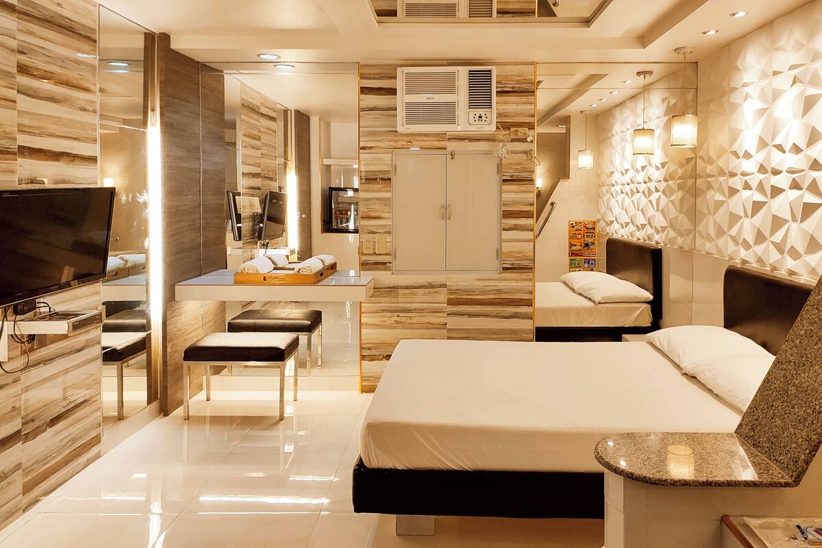 A modern hotel room with a large bed, a desk with a flat-screen TV, a seating area with a bench, and a decorative wall with geometric patterns.