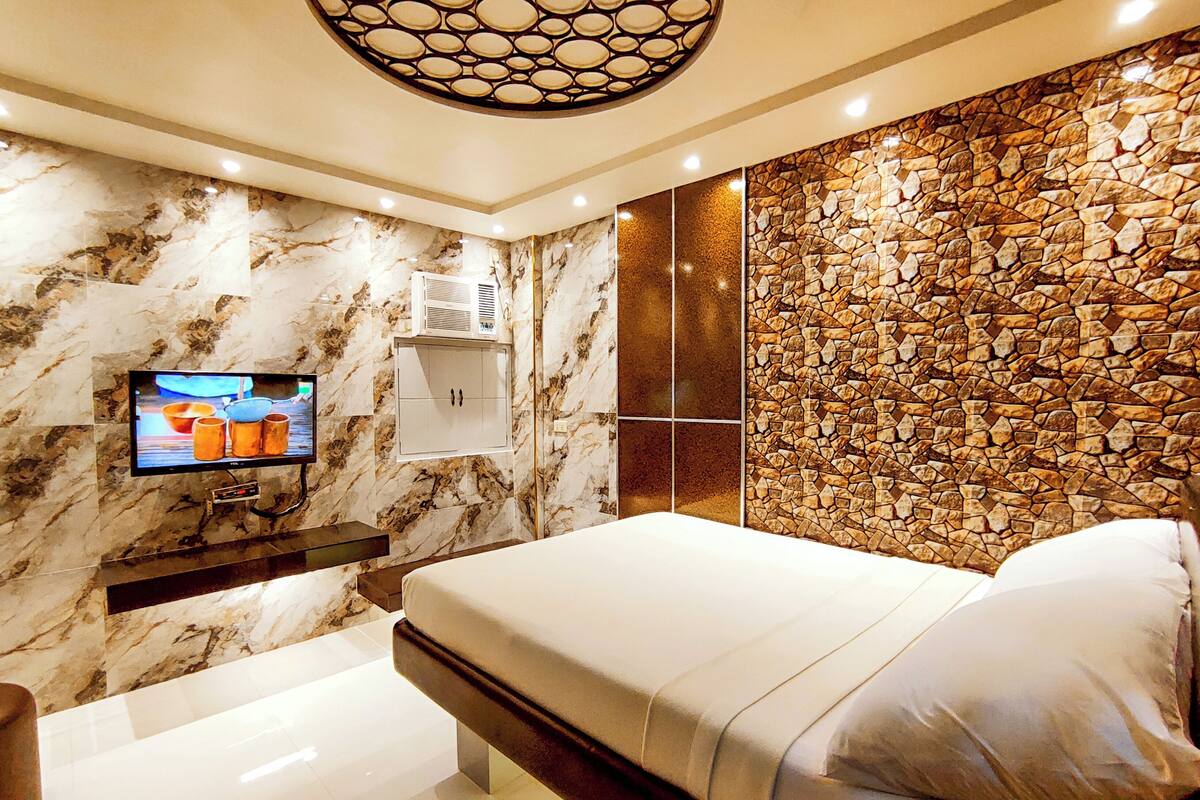 A modern bedroom with a marble wall, a stone accent wall, a flat-screen TV, a bedside table, and a bed with pillows.