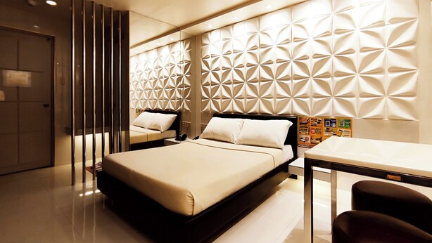 Una habitación de hotel moderna con una cama grande, un escritorio y una silla. La habitación tiene un muro con un patrón geométrico y una puerta con picaporte.