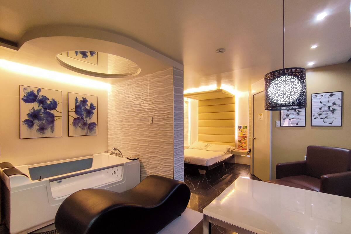 A modern spa room with a massage chair, a white bathtub, and a bed with a headboard.