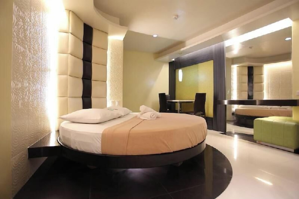 Una habitación de hotel moderna con una cama redonda, una mesita pequeña, un reposapiés verde y un espejo grande.