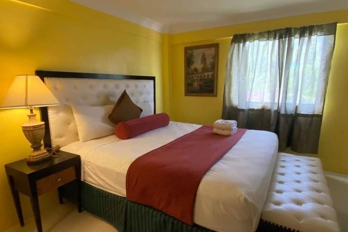 Une chambre d’hôtel avec un lit, une table de chevet, une lampe, une fenêtre avec des rideaux et un tableau au mur.