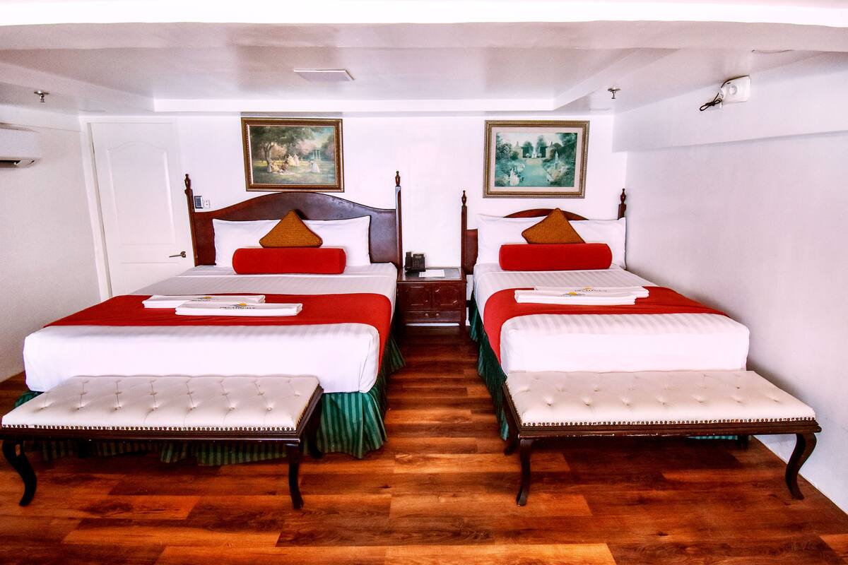 Une chambre d’hôtel avec deux lits, chacun recouvert d’une couvre-lit rouge et blanche, et dotés de têtes de lit assorties. On y trouve deux tables de chevet avec des lampes, un plancher en bois et une porte avec un cadre blanc.