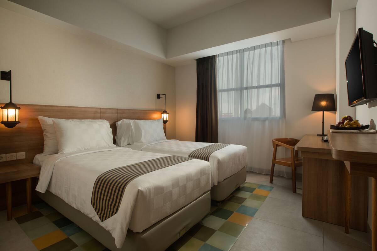 Ein Hotelzimmer mit zwei Betten, einem Schreibtisch, einem Stuhl, einem Fernseher und einem Fenster mit Vorhängen.