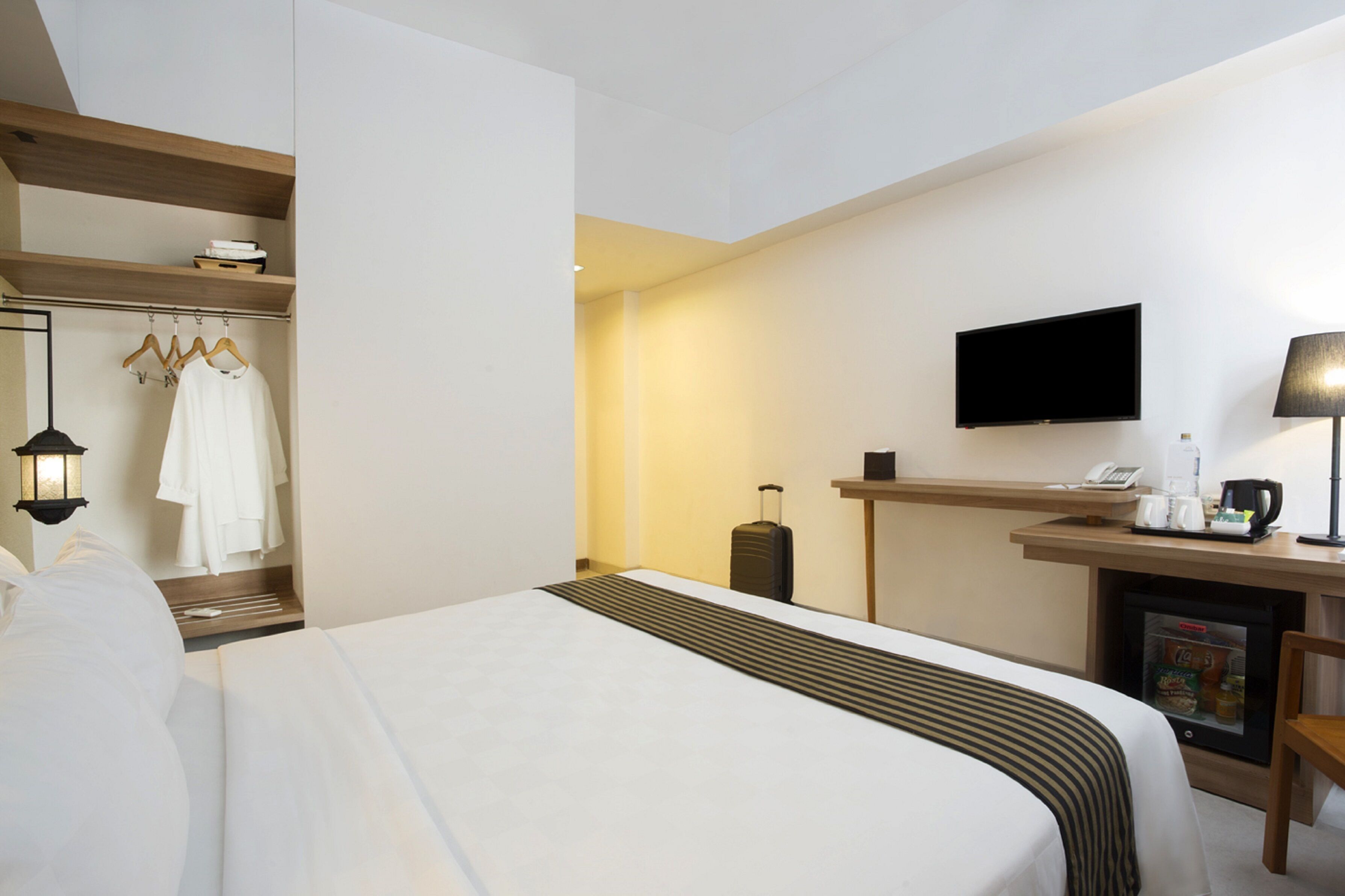 A hotel room with a bed, a desk with a flat-screen TV, a coffee maker, a mini-fridge, and a suitcase.