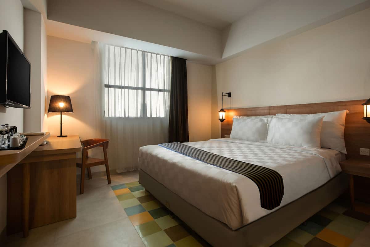Ein Hotelzimmer mit Bett, Nachttischlampen, einem Fernseher, einem Schreibtisch mit Stuhl und einem Fenster mit Vorhängen.