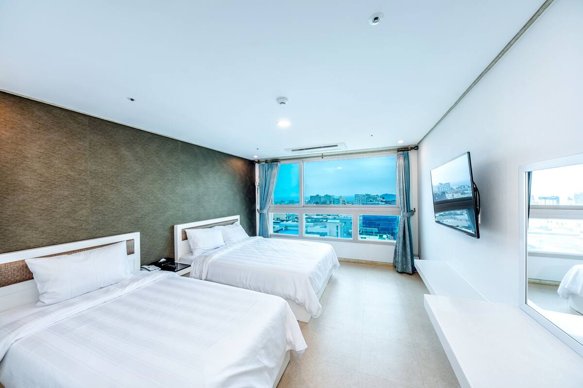 Ein modernes Hotelzimmer mit zwei Betten, einem großen Fenster mit Blick auf die Stadt, einem Flachbildfernseher und einer Bank am Fenster.