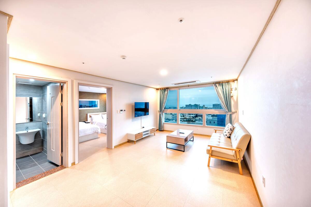 Ein modernes Wohnzimmer mit einer Couch, einem Couchtisch und einem Fernseher. Ein großes Fenster bietet einen Blick auf die Stadt.