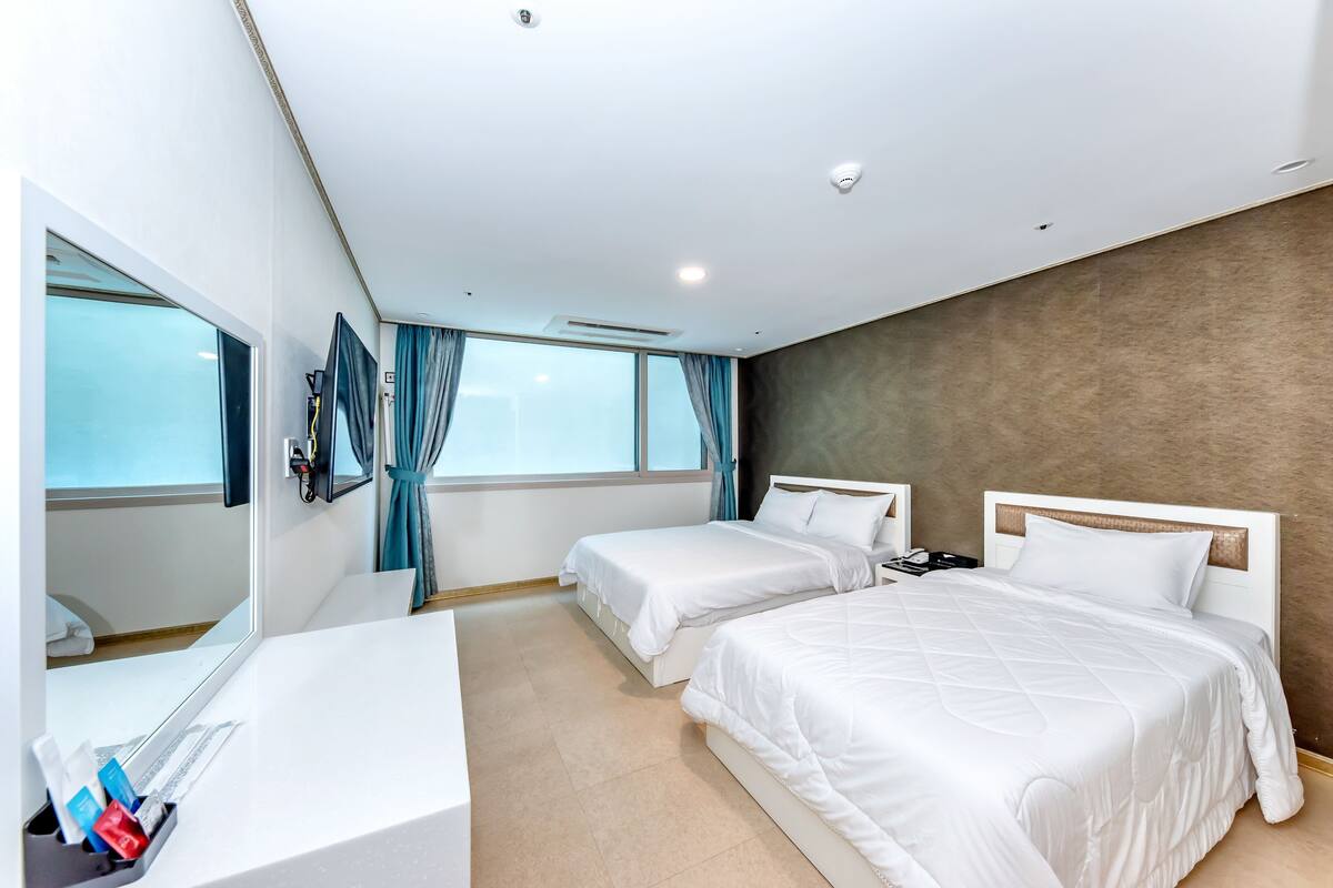 Ein Hotelzimmer mit zwei Betten, einem Schreibtisch, einem Spiegel und einem großen Fenster mit Vorhängen.