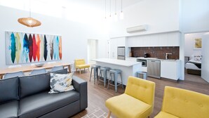 A modern living room with a gray sofa, yellow chairs, and a dining table. A kitchen with white cabinets and stainless steel appliances is visible in the background. - Loxton Courthouse Apartments (Loxton)