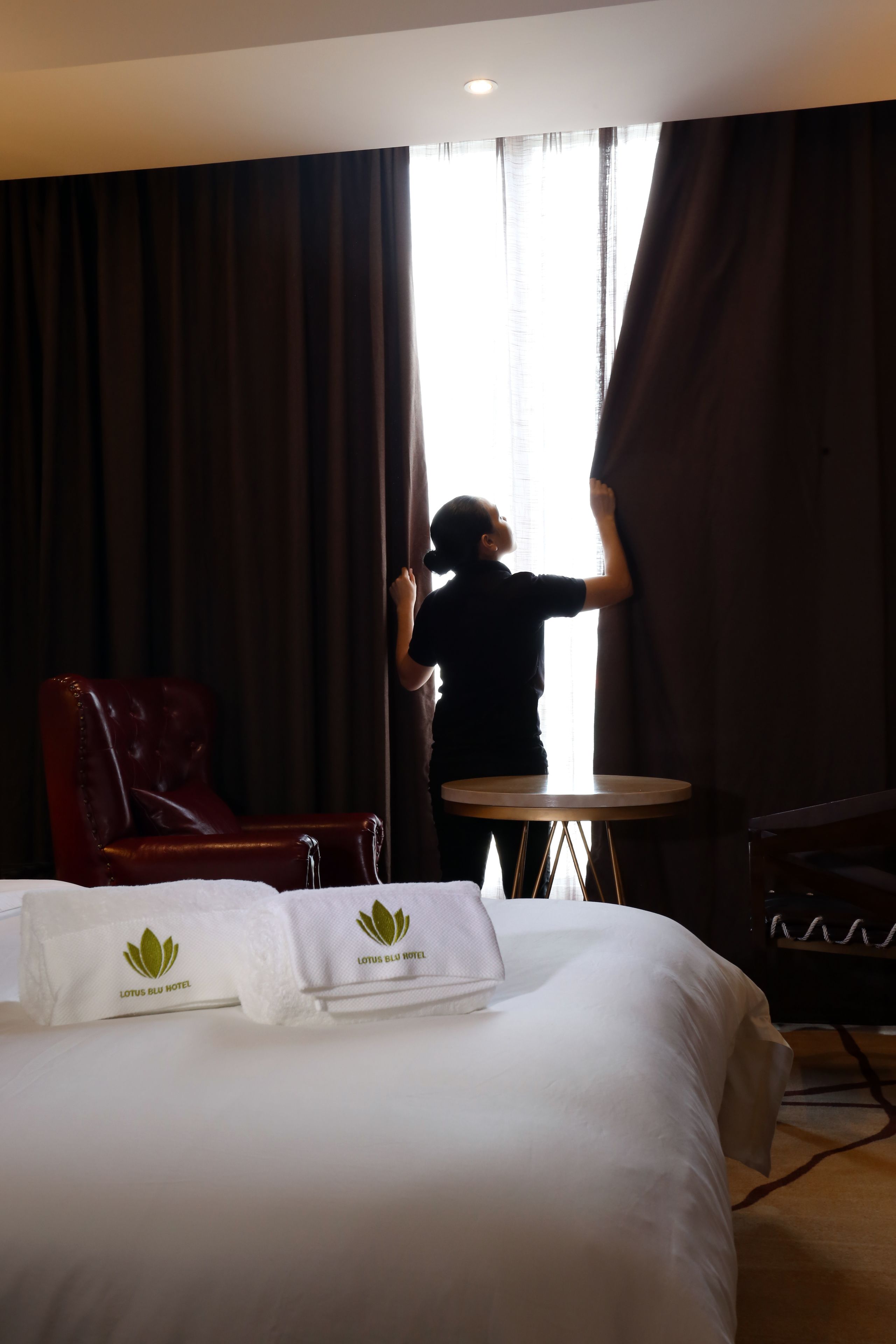 A hotel room with a bed, a red leather chair, a small round table, and a person adjusting curtains.