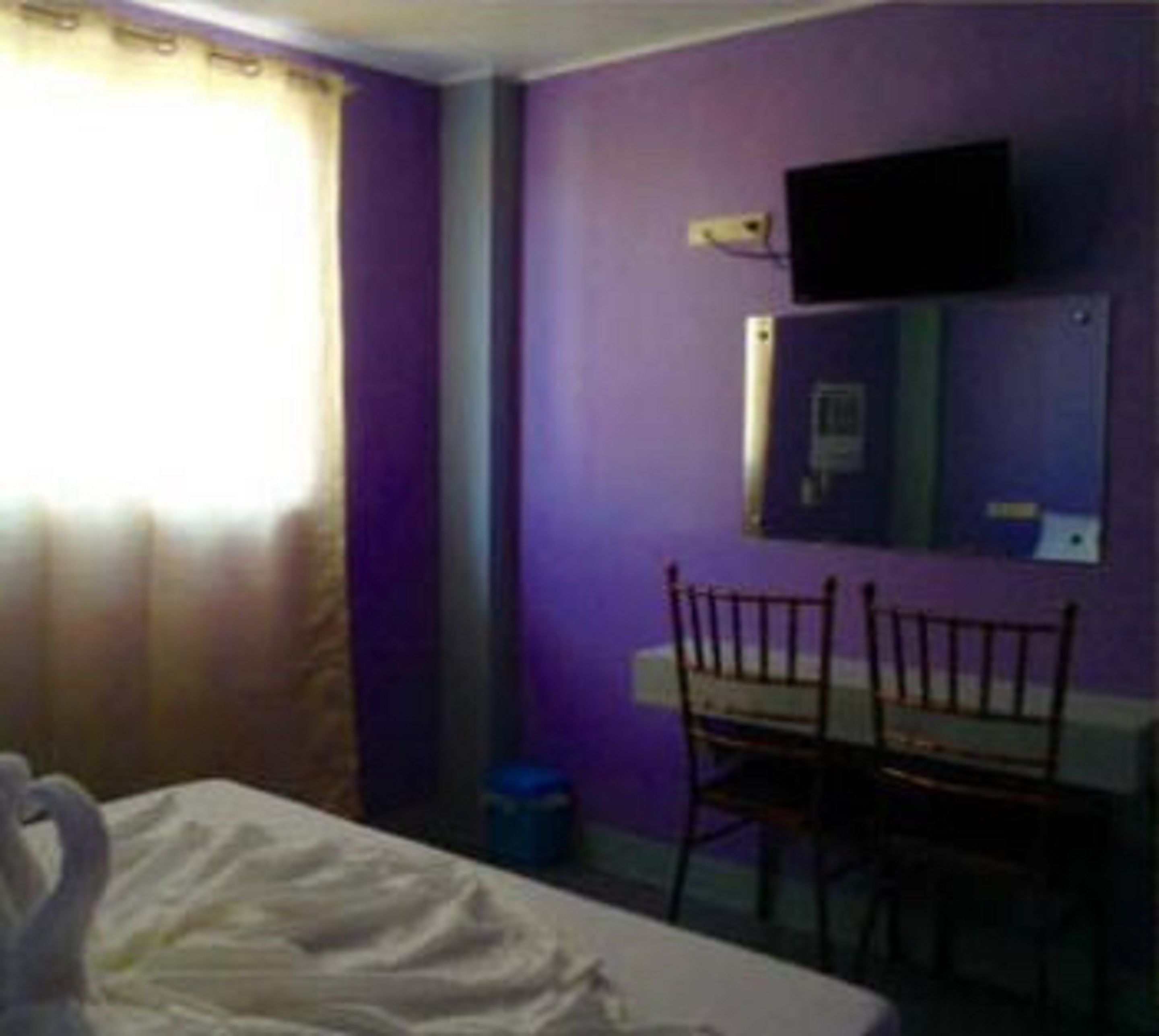 A room with a bed, a desk, and a chair. There is a window with curtains, a wall-mounted TV, and a mirror.