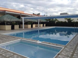 A swimming pool with a tiled deck, thatched umbrellas, and a building with a flat roof in the background.