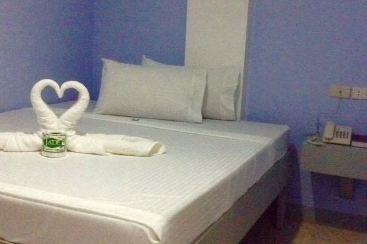 Una habitación de hotel con cama, almohadas blancas, una toalla dispuesta en forma de corazón, una mesita de noche pequeña con un teléfono y un enchufe de pared.