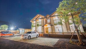 A two-story building with a well-lit entrance and a parking area with several cars. - Red Windmill Pension (Jeju City)