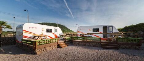 Two RVs parked on a gravel surface with picnic tables and a stone wall.