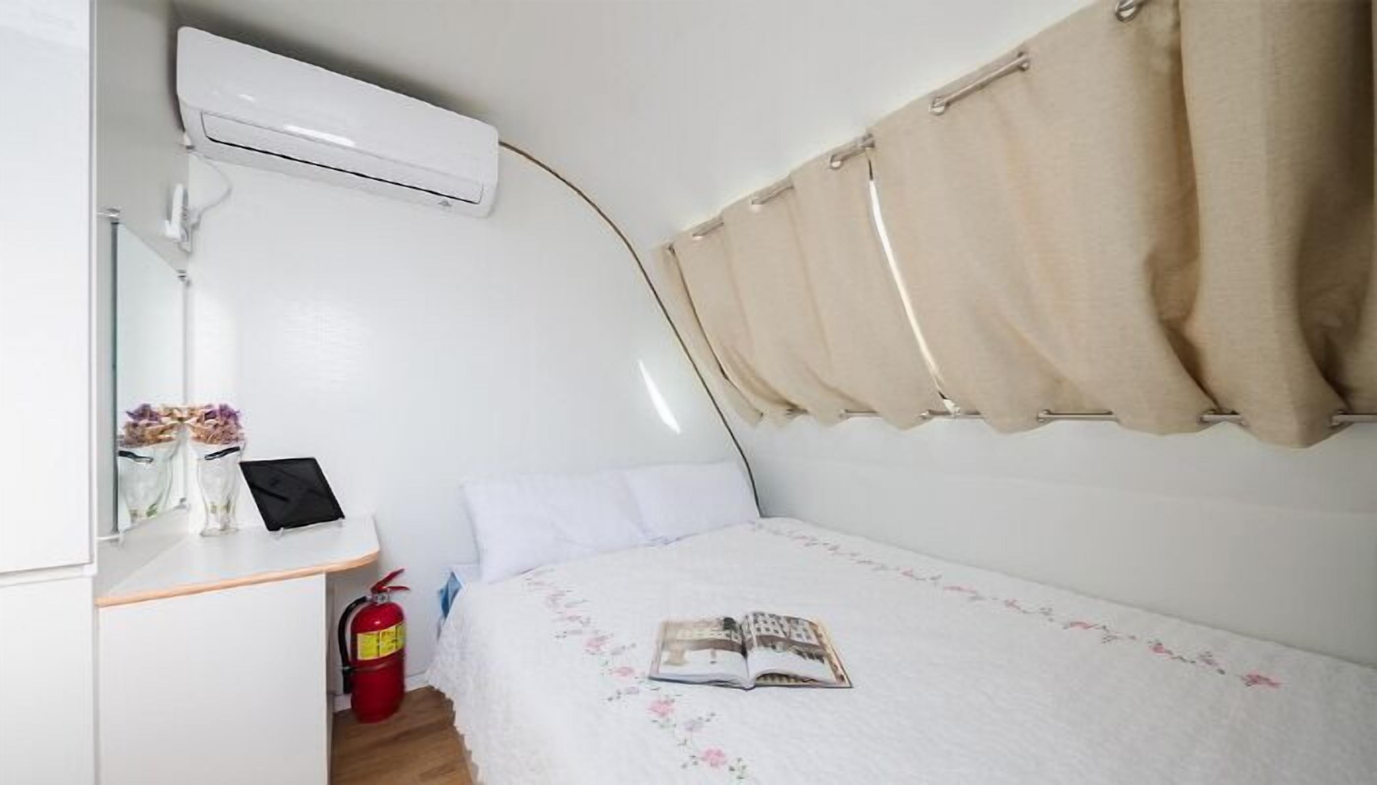 Een kleine, compacte kamer met een bed, nachtkastje met een lamp, brandblusser en raam met gordijnen.